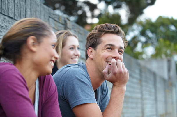 Дружеский разговор между друзьями. Снимок парня и двух девушек, которые вместе смеются, сидя на улице - Фото, изображение
