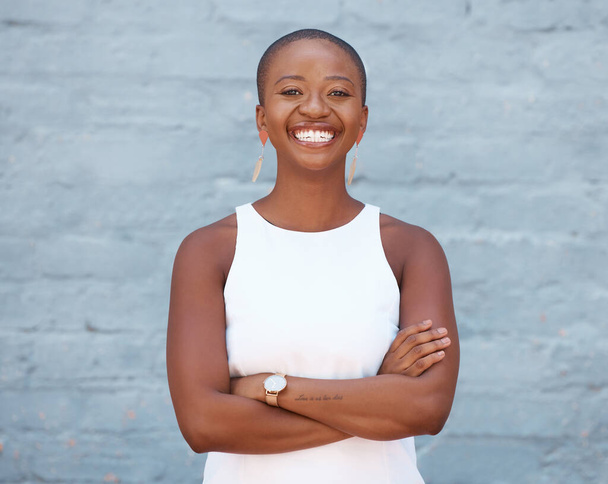Χαρούμενη φαλακρή Αφροαμερικανή που δείχνει σίγουρη ενώ στέκεται με σταυρωμένα τα χέρια έξω στον τοίχο. Ξέγνοιαστη νεαρή γυναίκα χαμογελά κοιτάζοντας την κάμερα. - Φωτογραφία, εικόνα