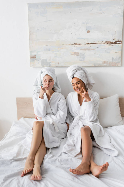full length of cheerful interracial women in white bathrobes and towels smiling and grimacing on bed at home - Foto, immagini
