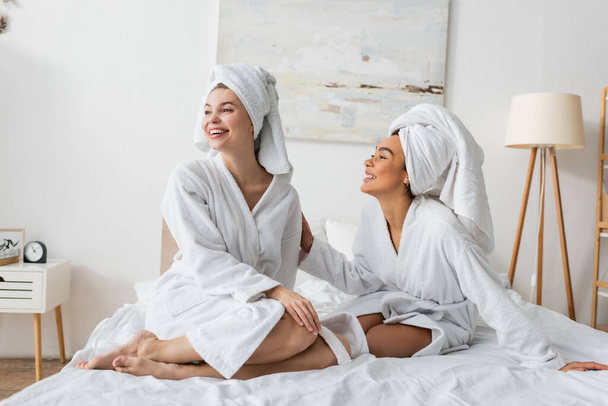 Ευτυχισμένη νεαρή γυναίκα με το μπουρνούζι και την πετσέτα να κοιτάζει μακριά κοντά στον χαμογελαστό Αφροαμερικανό φίλο της στο κρεβάτι. - Φωτογραφία, εικόνα