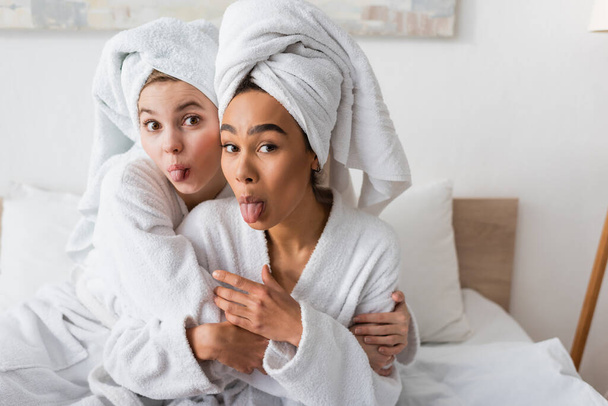 jonge interraciale vrouwen in witte handdoeken en badjassen kijken naar de camera en steken tongen in de slaapkamer - Foto, afbeelding