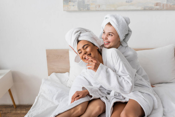 opgewonden Afrikaans amerikaanse vrouw in witte badjas en handdoek lachen met gesloten ogen in de buurt gelukkig vriend op bed - Foto, afbeelding