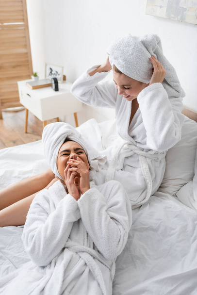 excited african american woman covering mouth while laughing near friend adjusting white towel on head - Valokuva, kuva