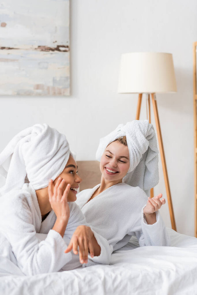 joyful interracial women in white bathrobes and towels gesturing during conversation in bedroom - Foto, Bild