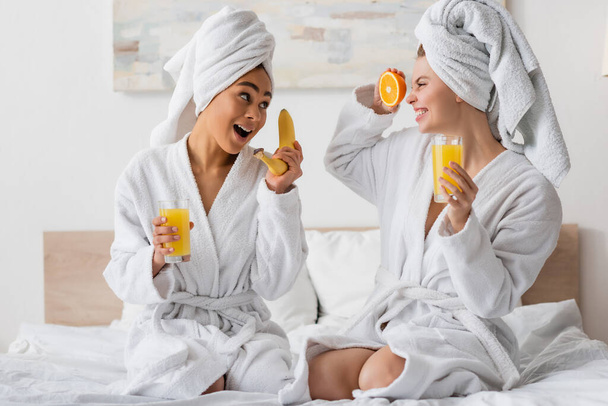 興奮した多民族女性で新鮮な果物やオレンジジュースを楽しみながらベッドに座って - 写真・画像