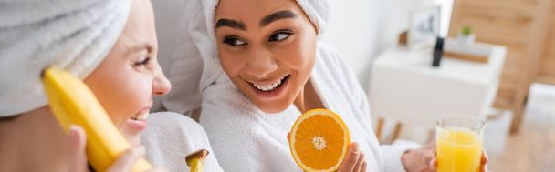 joyful african american woman holding fresh orange and juice near blurred friend with ripe banana, banner - 写真・画像