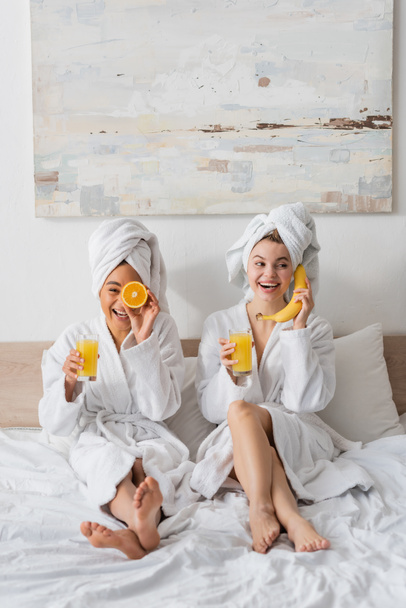 longitud completa de alegres mujeres interracial con frutas y jugo de naranja divertirse mientras se sienta en la cama - Foto, imagen