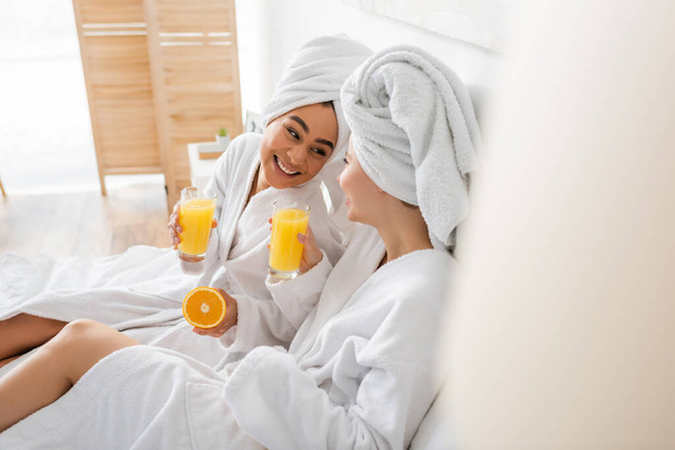 joyful african american woman with ripe orange and juice looking at friend in bedroom on blurred foreground  - Fotografie, Obrázek