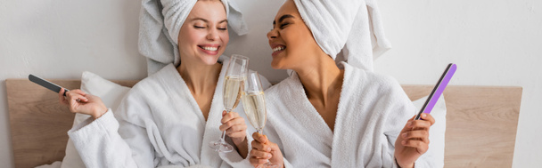 pleased multiethnic women in terry bathrobes and towels holding nail files and clinking champagne glasses in bedroom, banner - Photo, Image