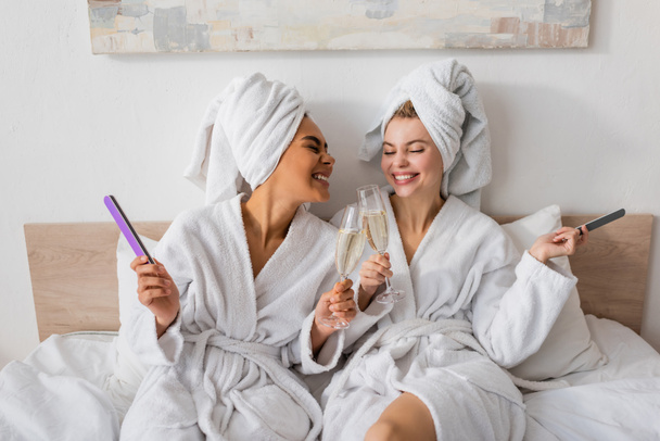 joyful multiethnic women in white terry robes and towels holding nail files and clinking champagne glasses on bed - Photo, Image