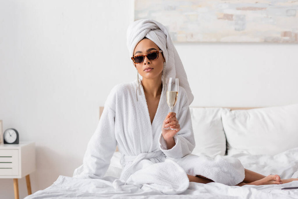 Barfüßige Afroamerikanerin in weißem weichen Handtuch und stylischer Sonnenbrille mit Champagnerglas auf dem Bett sitzend - Foto, Bild