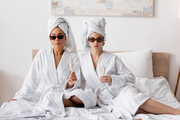 νεαρές πολυεθνικές γυναίκες με λευκά μπουρνούζια και γυαλιά ηλίου που κάθονται με ποτήρια σαμπάνιας και κοιτάζουν την κάμερα στην κρεβατοκάμαρα - Φωτογραφία, εικόνα