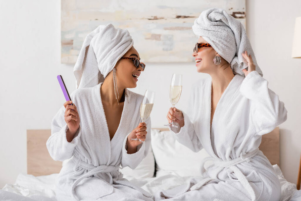 Ευτυχισμένες διαφυλετικές γυναίκες με μπουρνούζια και κομψά γυαλιά ηλίου κρατώντας σαμπάνια και κοιτώντας η μία την άλλη στην κρεβατοκάμαρα - Φωτογραφία, εικόνα