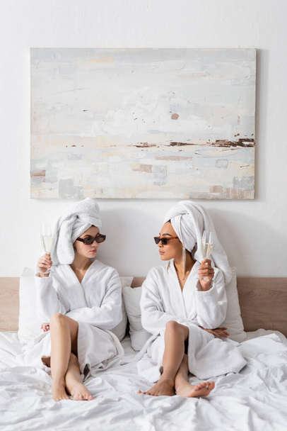 full length of barefoot interracial women in bathrobes and trendy sunglasses holding champagne and looking at each other on bed - Foto, Imagem