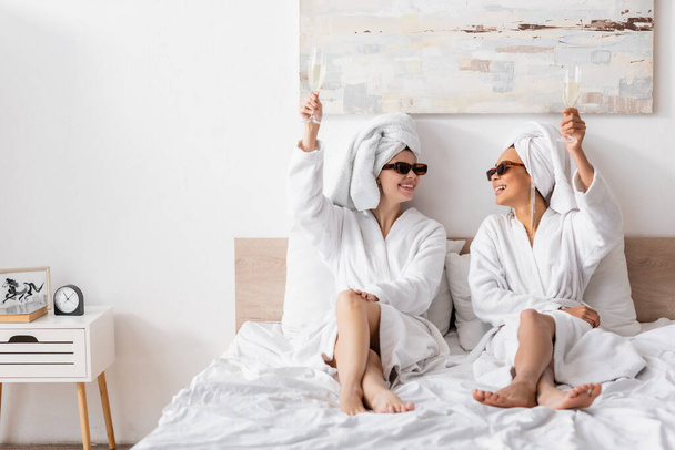 full length of joyful multiethnic women in terry bathrobes and sunglasses sitting on bed and toasting with champagne - Foto, Imagem