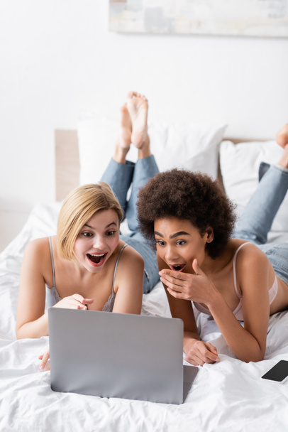 amazed interracial women in lingerie and jeans lying on bed and looking at laptop - Foto, Bild