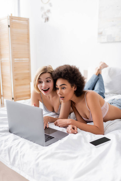 изумленная блондинка, указывающая на ноутбук рядом с африканским американским другом и смартфоном с пустым экраном на кровати - Фото, изображение