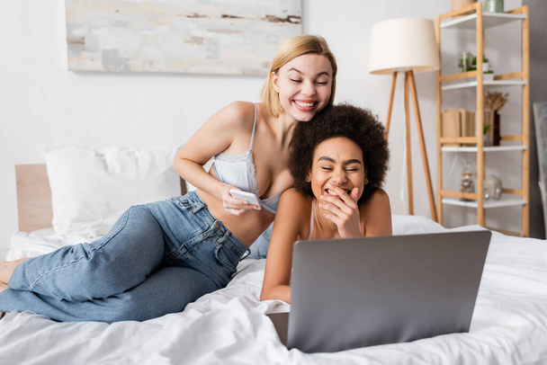 excited multiethnic friends in lingerie and jeans watching film on laptop in bedroom - Photo, Image