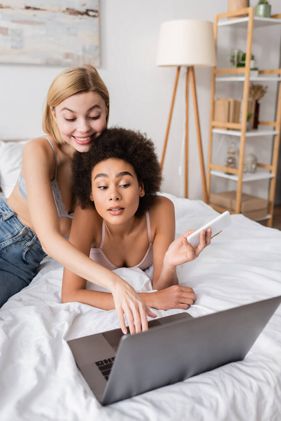 african american woman holding smartphone near smiling blonde friend using laptop on bed - Photo, Image