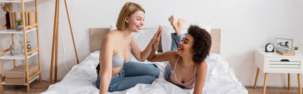 Modern yatak odasında sutyen ve kot pantolonlu mutlu ırklar arası kadınlar beşlik çakıyor. - Fotoğraf, Görsel