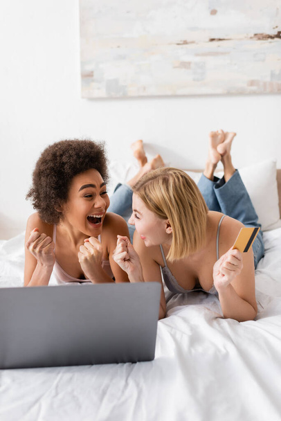 amigos multiétnicos emocionados mirándose entre sí y mostrando el gesto de ganar cerca de la tarjeta de crédito y el ordenador portátil en la cama en casa - Foto, imagen