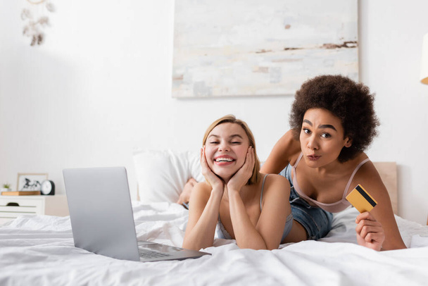african american woman holding credit card and grimacing near cheerful blonde friend and laptop on bed  - Photo, Image