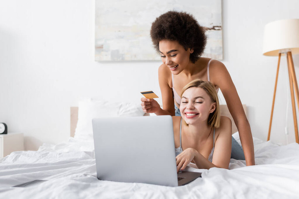 mujer afroamericana feliz sosteniendo la tarjeta de crédito cerca de amigo sonriente usando el ordenador portátil en la cama - Foto, imagen