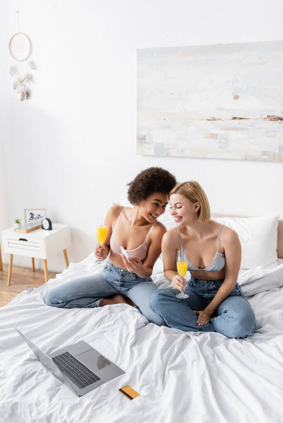 happy african american woman showing smartphone to smiling friend while sitting with cocktail near laptop and credit card on bed - Zdjęcie, obraz