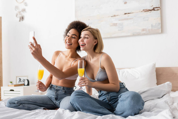 young and happy interracial women with cocktails in champagne glasses sitting on bed and taking selfie on cellphone - Foto, imagen