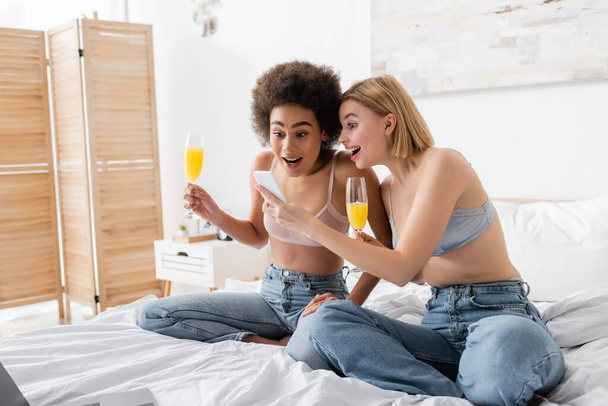amazed interracial women in lingerie and jeans holding cocktails and looking at smartphone in bedroom - Zdjęcie, obraz