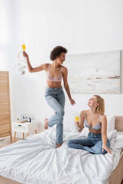 joyful african american woman in bra and jeans posing with cocktail near blonde friend sitting on bed - Foto, imagen
