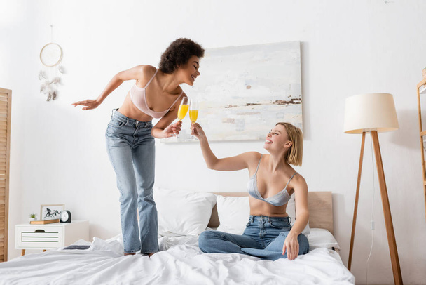 joyful interracial women in jeans and bras clinking champagne glasses with cocktails in bedroom - Photo, Image