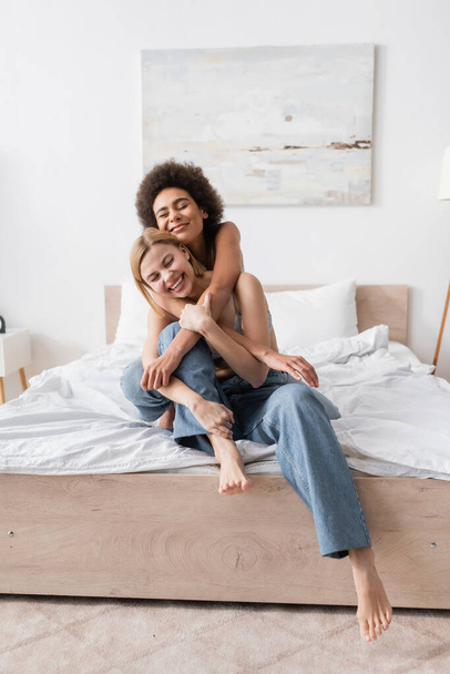 happy african american woman embracing blonde barefoot friend in jeans sitting on bed at home - Zdjęcie, obraz