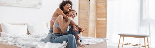 joyful african american woman embracing blonde friend sitting on bed in bra and jeans, banner - Foto, Imagem