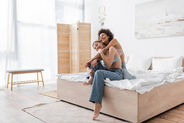 cheerful african american woman in bra and jeans hugging happy friend in modern spacious bedroom - Photo, Image