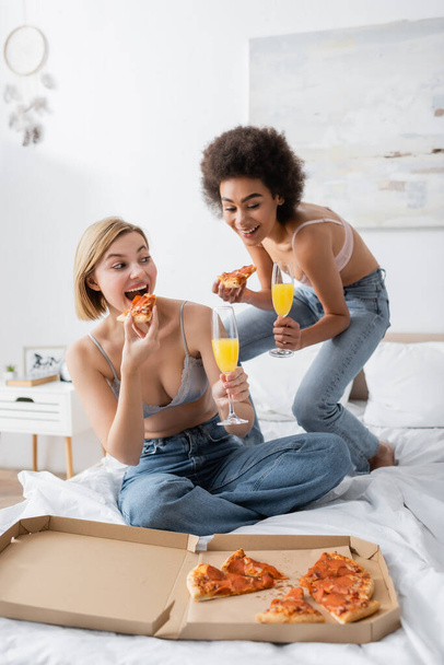 χαρούμενη Αφροαμερικανή γυναίκα κρατώντας κοκτέιλ κοντά σε ξανθιά φίλη τρώγοντας πίτσα στο κρεβάτι στο σπίτι - Φωτογραφία, εικόνα