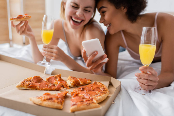 excited multiethnic women with cocktails laughing near smartphone and tasty pizza in bedroom - Foto, Imagen