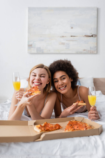 cheerful interracial friends with tasty pizza and cocktails in champagne glasses lying on bed and looking at camera - Zdjęcie, obraz
