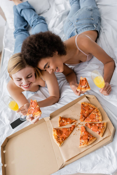 visão de alto ângulo de mulheres inter-raciais sorrindo com olhos fechados perto de pizza saborosa e coquetéis em copos de champanhe enquanto deitado na cama - Foto, Imagem