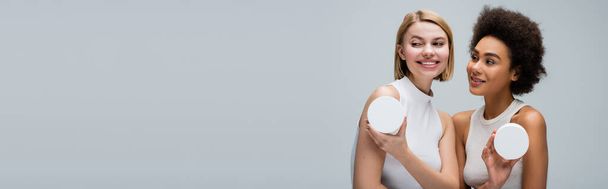 pretty african american woman looking at blonde model posing with container of body cream isolated on grey, banner - Foto, Imagen
