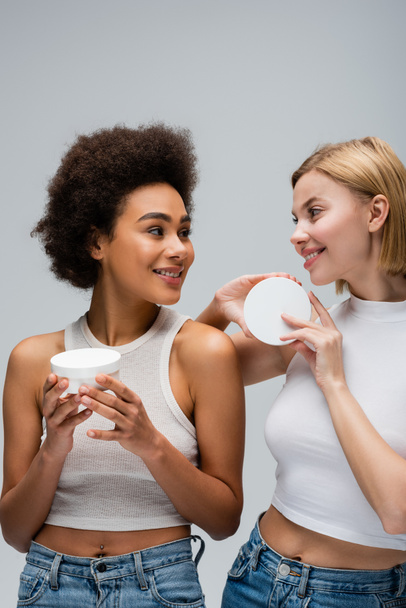pretty interracial women in white tops holding containers with cosmetic cream and smiling at each other isolated on grey - Foto, imagen