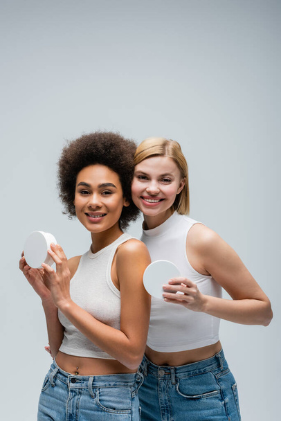 jolis modèles multiethniques en débardeurs blancs posant avec des récipients de crème cosmétique isolés sur gris - Photo, image