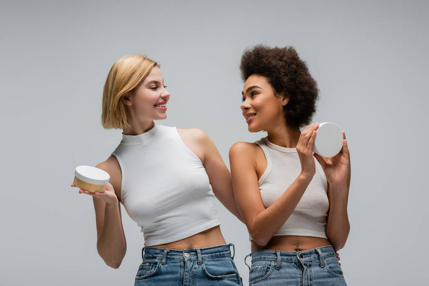 young multiethnic women holding body cream and smiling at each other isolated on grey - Foto, Bild