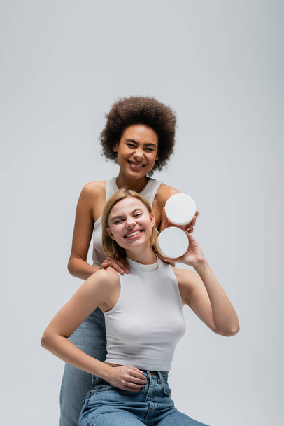 excited multiethnic models in white tops and jeans posing with body cream and looking at camera isolated on grey - Photo, image
