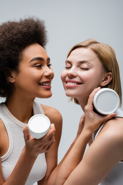happy blonde model with closed eyes posing with cosmetic cream near african american woman isolated on grey - Fotó, kép