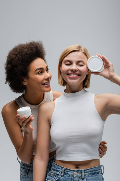 happy blonde model with closed eyes posing with body cream near african american woman isolated on grey - Foto, Bild