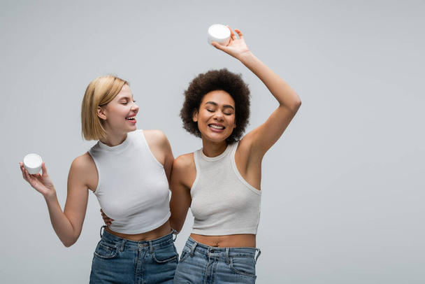 cheerful african american model posing with body cream in raised hand near blonde woman isolated on grey - Fotografie, Obrázek
