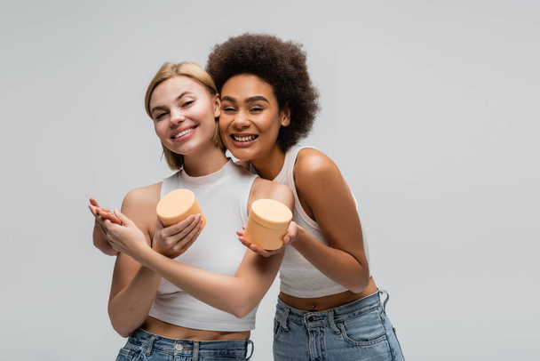 pleased multiethnic women holding hands while posing with cosmetic cream isolated on grey - Fotó, kép