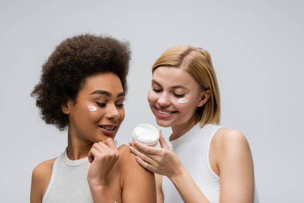 pleased multiethnic women in white tops looking at container with face cream isolated on grey - Zdjęcie, obraz