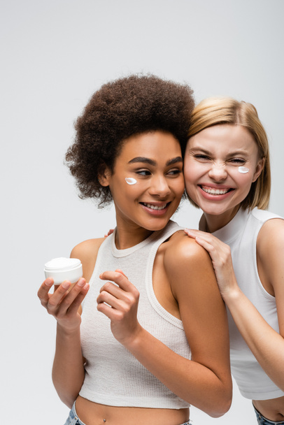 cheerful blonde woman looking at camera near african american model posing with face cream isolated on grey - Foto, immagini